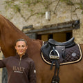 Woman standing next to a horse with a saddle, wearing a brown jacket with 'HALT' branding.